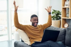 man sitting on a couch cheering, he is happy while looking at his laptop