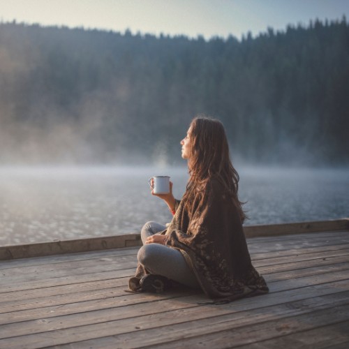 woman sitting in front of a lake, holding a cup of coffee