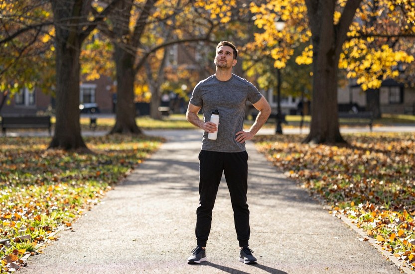 Man in his mid thirties pausing on a running path in a US city park in early morning autumn light, water bottle in hand, the kind of run people count on to detox from weed at home